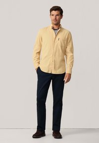 Camisa de botones amarillo claro con un bolsillo, superpuesta a una camisa a rayas, combinada con pantalones navy oscuros y zapatos marrones.