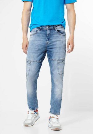 Person wearing blue faded jeans, colorful sneakers, and a bright blue t-shirt, standing against a white background.