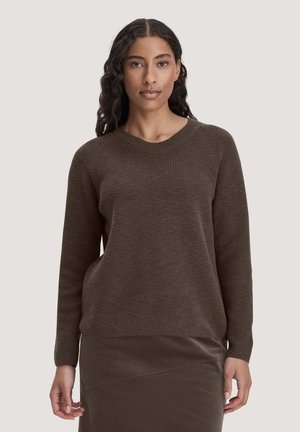 Femme aux longs cheveux bouclés et foncés, portant un pull en maille marron et une jupe marron assortie, debout devant un fond uni clair.