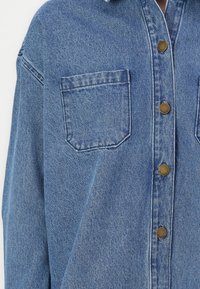 Camicia di denim azzurro chiaro, caratterizzata da un colletto a punta, due tasche sul petto e bottoni metallici marroni. Texture strappata evidente.