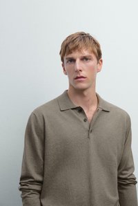 Jonge man met lichtbruin haar die een bruin poloshirt met lange mouwen draagt, staande tegen een effen lichtgrijze achtergrond.