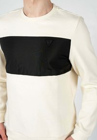 Sudadera de cuello redondo en marfil y negro, con un diseño de bloques de color horizontal, textura suave y un pequeño logotipo triangular en el pecho.