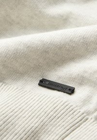 Tissu tricoté gris clair avec une texture subtile. Une petite étiquette noire avec "STRAIGHTONE" est visible, portant un design texturé et une forme rectangulaire.