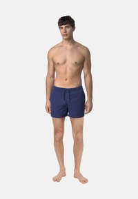 Pantaloni da bagno blu navy con vita regolabile, realizzati in tessuto leggero. Caratterizzati da una vestibilità rilassata e tasche laterali. I piedi nudi, in posa eretta.