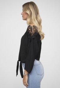 Svart topp med båret skulderparti med blondekanter, lange ermer og knyttet midje. Kombinert med lyseblå jeans. Jevn stofftekstur.