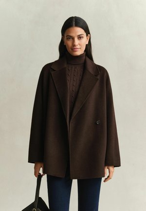 Femme debout face à l'objectif, portant un manteau marron foncé sur un pull à col roulé et un jean bleu, tenant un sac à main noir dans sa main gauche.
