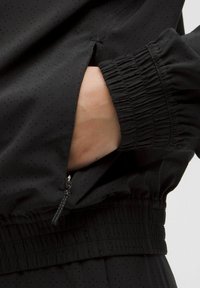Čierna ľahká bunda s prístupnou kapsou na zips, elastickými manžetami a perforovanou textúrou pre lepšiu priedušnosť.