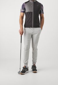 Golfové tričko v černé, šedé a vzorovaných rukávech, kombinované se světle šedými kalhotami. Oblečeno s tmavými teniskami, držící golfovou hůl.