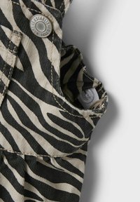 Zebra-mönstrad tyg i svart och beige, med en metallknapp och synliga sömnadsdetaljer. Totalt sett ett texturerat utseende och känsla.