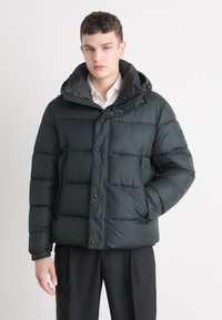 Grønn pufferjakke med hette, med horisontal quilting, trykknapper og sidelommer. Laget av et glatt syntetisk materiale.