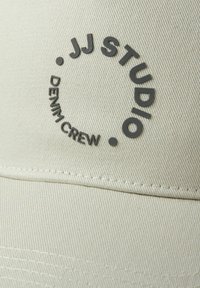 Beige cap with a structured design, featuring a circular logo in dark gray text. The fabric has a textured, ribbed appearance.