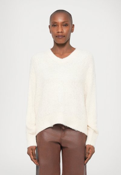 White, textured knit sweater with a V-neck, dropped shoulders, and a high-low hem. Worn with brown, shiny leather pants.