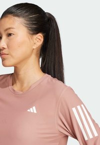 Světle růžové sportovní tričko s texturovanou tkaninou. Obsahuje bílé logo a tři pruhy na levém rukávu. Design s kulatým výstřihem.