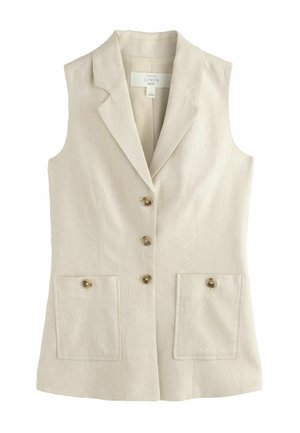 Femme marchant vers l'avant portant un blazer beige sans manches et un pantalon droit assorti avec des sandales sur un fond clair.