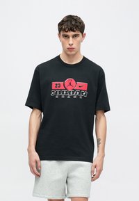 Μαύρο βαμβακερό μπλουζάκι με γραφικό σχέδιο σε κόκκινο και λευκό με την επωνυμία "JORDAN". Κοντά μανίκια και άνετη εφαρμογή, συνδυασμένο με γκρι σορτς.