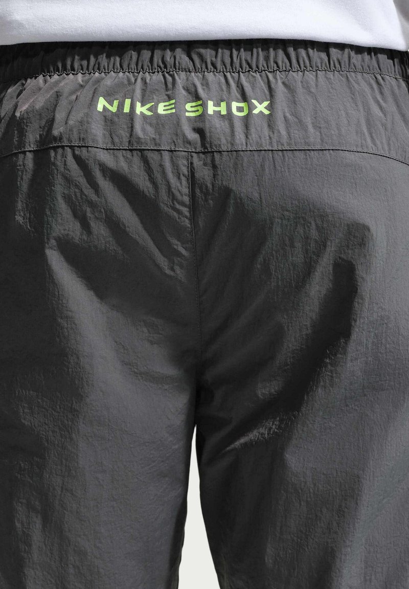 Persoon draagt zwarte Nike Shox broek, elastische tailleband zichtbaar met lichtgroen logo op de achterkant onder een wit shirt.