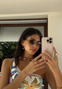 Akiniai nuo saulės su žaliai tonuotais lęšiais, balta raštuota palaidinė, keli sluoksniuoti vėriniai ir daug žiedų iš įvairių metalų ir akmenų.