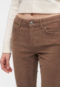 Femme portant un pantalon en velours côtelé marron avec bouton et fermeture éclair, associé à un pull court en maille torsadée crème, main détendue sur le côté.