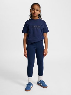 Pige stående med afslappede hænder, iført navyblåt sportstøj, hvide sokker og blå sneakers mod en ensfarvet baggrund.