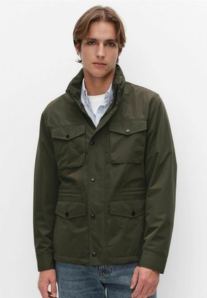 Jonge man die een donkergroene jas met vier voorzakken draagt, over een lichtblauw overhemd en een wit ondershirt, tegen een effen achtergrond.