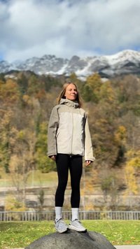 Világos bézs és szürke kapucnis dzseki, fekete leggings, és szürke cipők. Egy sziklán áll természetes háttérrel, mely fákból és hegyekből áll.