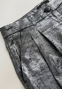 Pantaloni gri plisati dintr-un material strălucitor, texturat, cu închidere prin buton și etichetă de marcă. Prezintă variații tonale subtile.