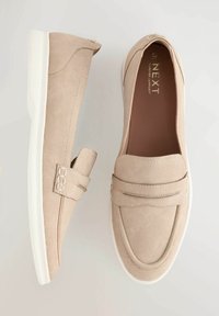 Beige suede slip-on shoes with a rounded toe, featuring a decorative strap and white rubber sole. Smooth texture, minimal stitching details.