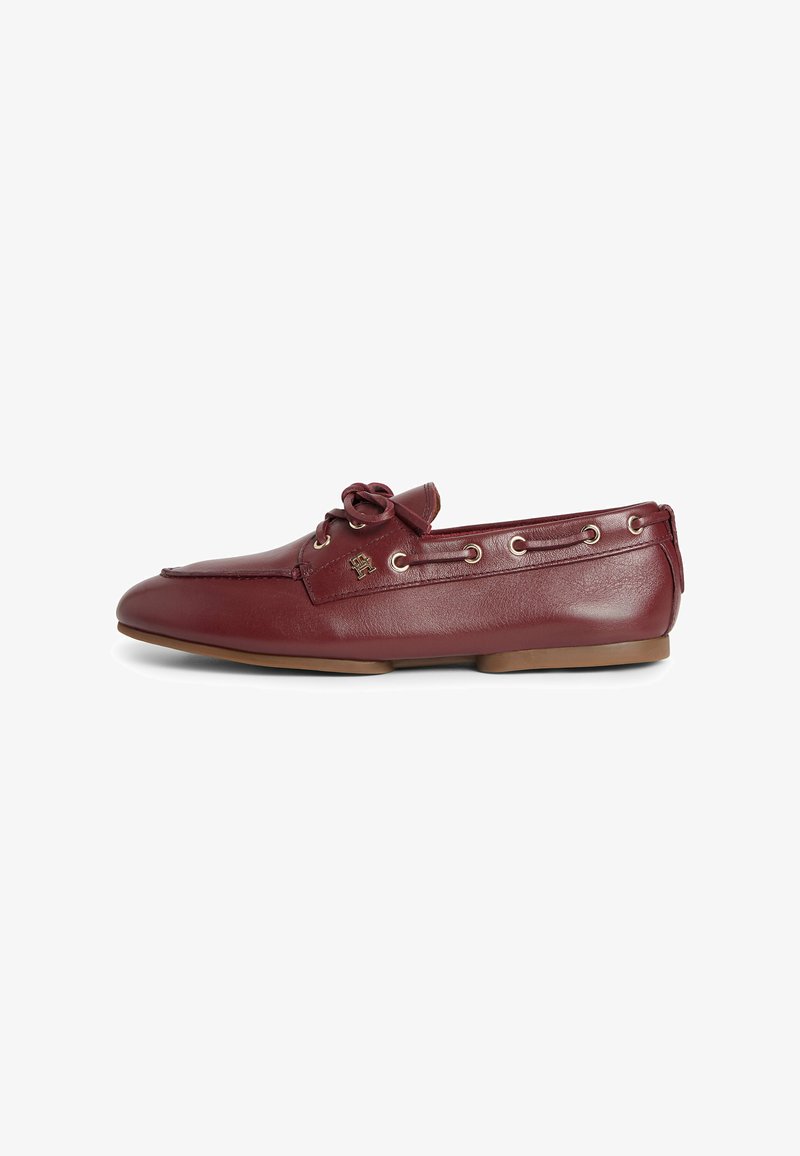 Mocassin en cuir bordeaux avec détail nœud, œillets argentés le long des côtés, semelle plate marron et petit logo en métal sur le dessus.