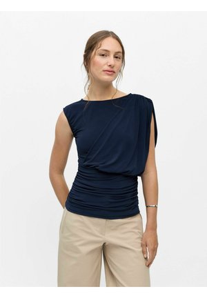 Frau mit heller Haut und zurückgebundenem Haar, die ein ärmelloses dunkelblaues Oberteil mit drapiertem Design und beige Hose vor einem einfarbigen weißen Hintergrund trägt.