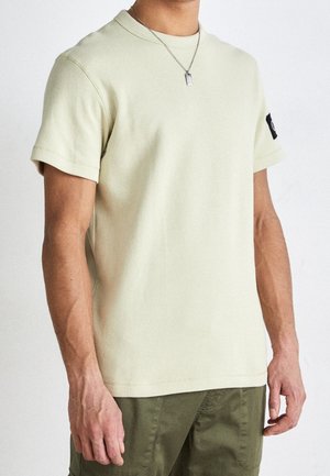 Un homme portant une chemise en tissu texturé beige clair à manches courtes, un pantalon vert olive et un collier avec un pendentif rectangulaire argenté.
