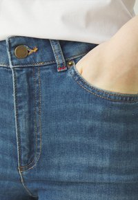 Jeans de mezclilla azul con un corte relajado, que presentan costuras en contraste, un cierre de botón metálico y un acento rojo en el forro del bolsillo.
