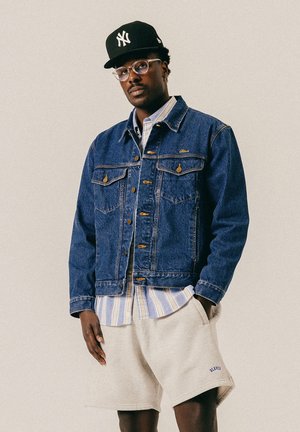 Mężczyzna w czarnej czapce New York Yankees, okularach, niebieskiej kurtce dżinsowej, pasiastym t-shircie i beżowych szortach, trzymający ręce w kieszeniach na jednolitym tle.