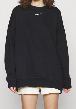 Persona con una sudadera negra de Nike de gran tamaño con logo blanco y pantalones cortos blancos, recortados a la mitad del muslo, contra un fondo liso.