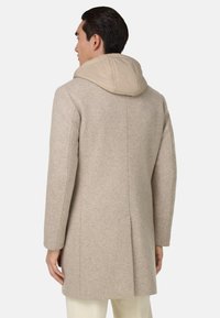 Béžový vlněný kabát s kontrastní hnědou vycpávanou kapucí, střih na míru a zadní rozparek; hladká textura s minimem švů.