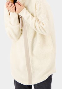 Persona che indossa una giacca a zip di pile color crema, con maniche lunghe e aperture per il pollice, mentre tiene il colletto vicino al viso.