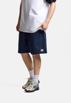 Mężczyzna ubrany w granatowe szorty z logo, białą koszulkę, białe skarpetki i beżowe tenisówki, stojący na jednolitym jasnym tle.