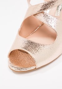 Metaliczne różowe złote buty typu open-toe z krzyżującymi się paskami, teksturowaną powierzchnią i gładkim wnętrzem. Płaska podeszwa z zaokrąglonym czubkiem.