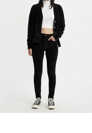 Femme portant un cardigan noir duveteux, un col roulé court blanc, un jean skinny noir et des baskets noires et blanches, debout sur fond blanc.