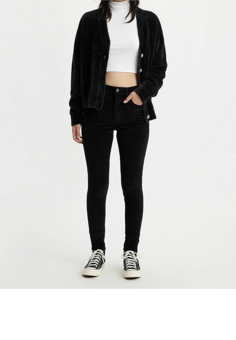 Femme portant un cardigan noir duveteux, un col roulé court blanc, un jean skinny noir et des baskets noires et blanches, debout sur fond blanc.