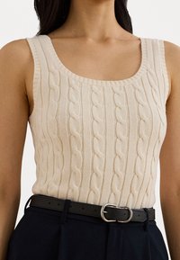 Beige stickad linne med kabelmönster, vid halsringning och ribbade kanter; parat med ett mörkt bälte med en silverbuckle.