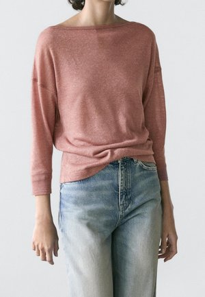 Personne portant un haut ample à manches longues rose poudré et un jean taille haute bleu clair, debout devant un fond uni.