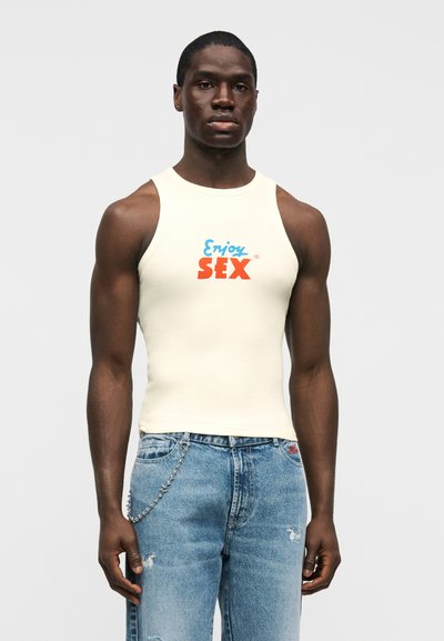 Εκρού φανελάκι με μπλε και κόκκινο κείμενο "Απόλαυσε το SEX", συνδυασμένο με μπλε τζιν με φθορές και ασημένιο αλυσίδα ως αξεσουάρ.