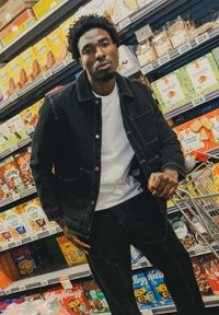 Veste en denim noir avec des coutures blanches, poches avant, associée à un t-shirt blanc et un pantalon noir. Présentée dans une allée de supermarché.