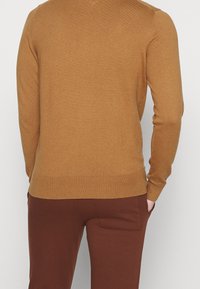 Brun stickad tröja med långa ärmar och rund halsringning. Texturen är slät och har en åtsittande design vid nederkanten. Kombinerad med mörkbruna byxor.