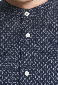 Camicia blu navy con motivo a rombi bianchi, colletto a fascia e quattro bottoni bianchi davanti. Tessuto morbido.