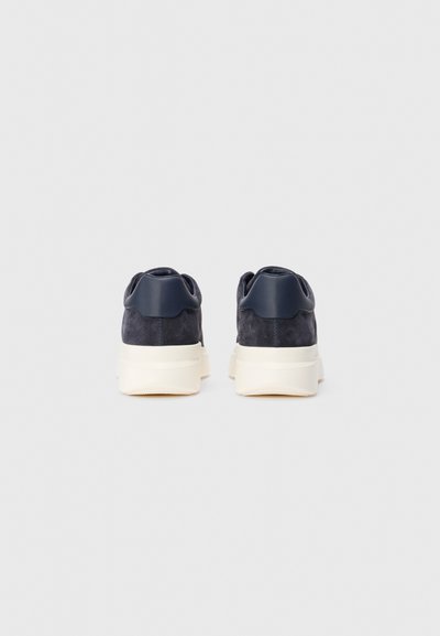 Pair of dark blue suede sneakers with white thick soles viewed from the back on a plain light background.