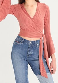 Dusty rose wraps-top, jossa on ribbed-tekstuuri ja kellotElems, yhdistettynä keskiharmaisiin korkeavyötäröisiin farkkuihin, joissa on klassinen viiden taskun muotoilu.