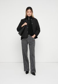 Gequiltte zwarte jas, zwarte coltrui, grijs panterprint broek, en puntige zwarte schoenen. Model houdt een zwarte handtas vast. Eenvoudige achtergrond.
