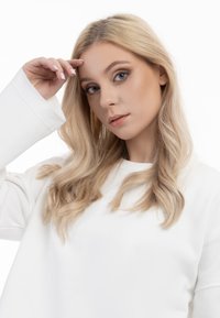 Sweatshirt bianco con un ampio scollo a giro e maniche lunghe e larghe. Capelli chiari con morbide onde incorniciano il viso, che presenta un trucco naturale.