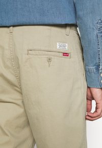 Beige chinos med en bakficka, med en Levi's etikett. Tyget verkar slätt, kompletterat med en ljusblå skjorta.